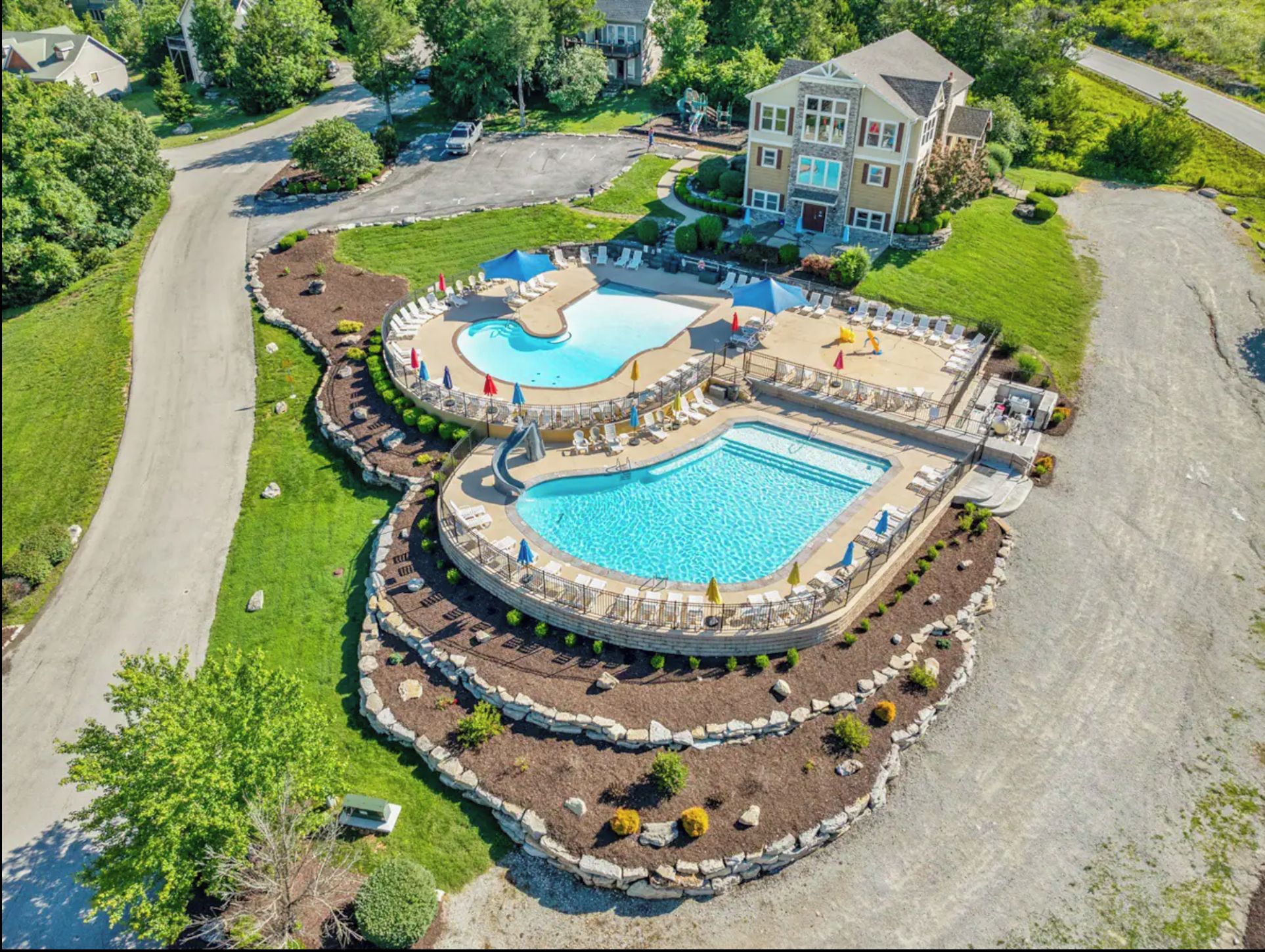 A large, multi-pool resort with a central swimming area surrounded by lounge chairs and umbrellas, set in a landscaped garden with a parking area and a large building in the background.