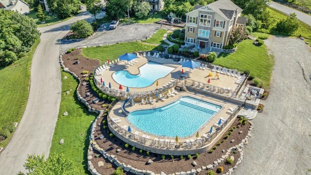 Ein großes Resort mit mehreren Pools, ein zentrales Schwimmbecken umgeben von Liegestühlen und Sonnenschirmen, eingebettet in einen angelegten Garten mit Parkplatz und einem großen Gebäude im Hintergrund.