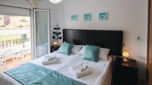 Ein ordentlich bezogenes Bett mit weißer und türkisblauer Bettwäsche, ein schwarzer Kopfteil und ein Balkon mit Stuhl und Tisch.