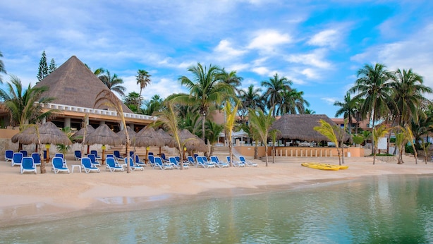 Un resort en la playa con estructuras de techo de paja, sillas de descanso azules y una piscina tranquila.