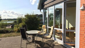 A patio with a table and chairs, a glass door leading to a room, and a view of a lake and buildings in the background. - 3 bedrooms to 6 people, Sauna, terrace, 4 * equipment, sea view (Sellin)