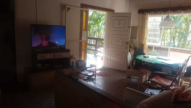 A living room with a television, a dining area with a table and chairs, and a view of the outdoors. - House 2 suites + 2 apartments - 12 people, Foot of the hill, nature at your window (Praia de Barequecaba)