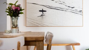 Une photographie encadrée représentant un surfeur sur une plage, une table à manger en bois et un vase rempli de fleurs.