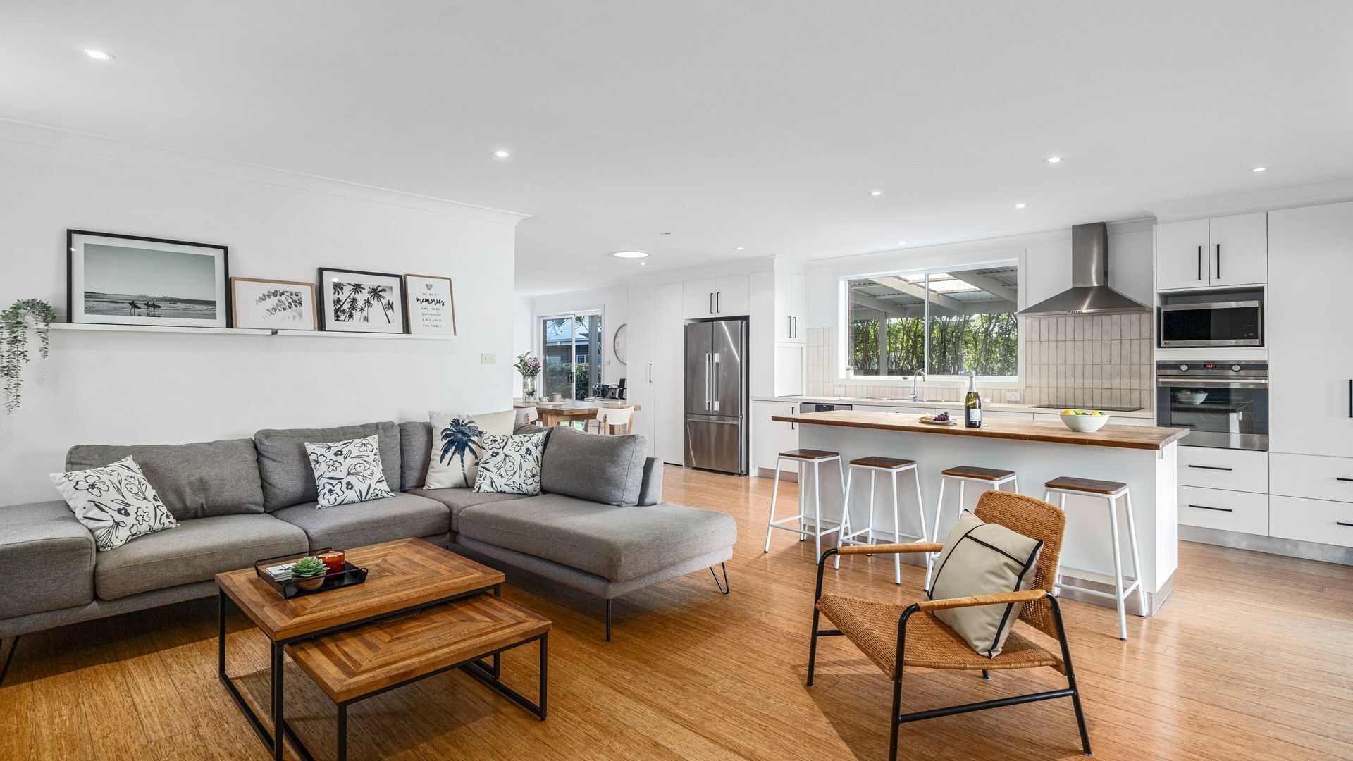 Ein modernes Wohnzimmer mit grauem Sofa, Couchtisch aus Holz und Korbstuhl. Im Hintergrund ist eine Küche mit weißen Schränken, einem Edelstahlkühlschrank und einer Bar erkennbar.