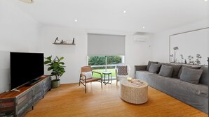 Un salon moderne avec un téléviseur à écran plat, une grande plante en pot, un canapé gris et une table basse en osier.