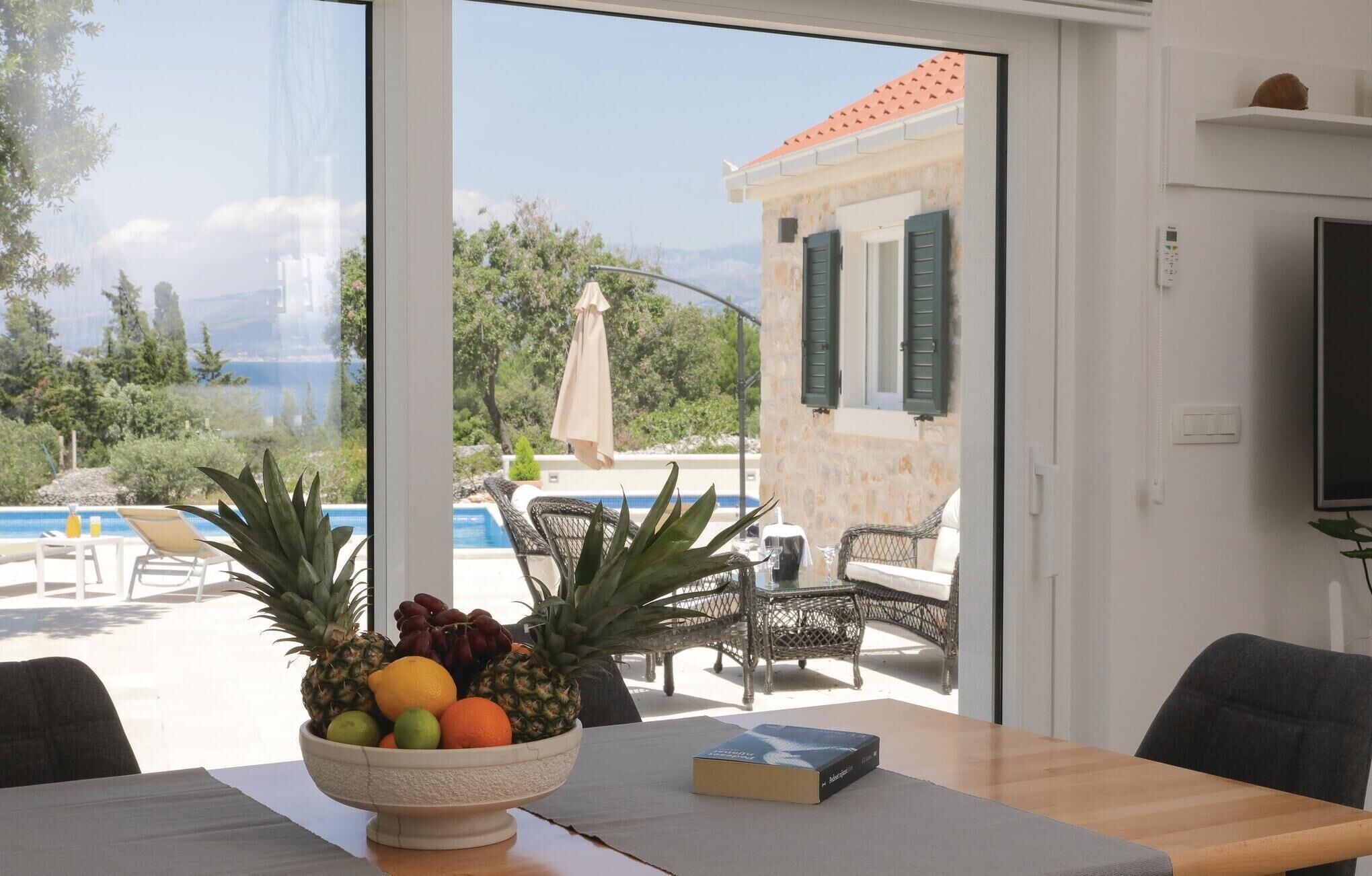 A room with a view of a pool and mountains, a table with a fruit bowl and a book, and a chair.