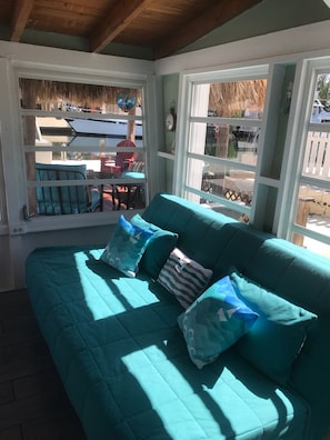 A teal-colored sofa with patterned cushions in a room with large windows, wooden beams, and a view of outdoor furniture.