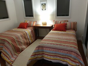 Ein Schlafzimmer mit zwei Betten, jeweils mit gemusterten Bettbezügen und roten Kissen. In der Mitte steht ein Holztisch mit einer Lampe darauf, darüber an der Wand ein gerahmtes Bild.