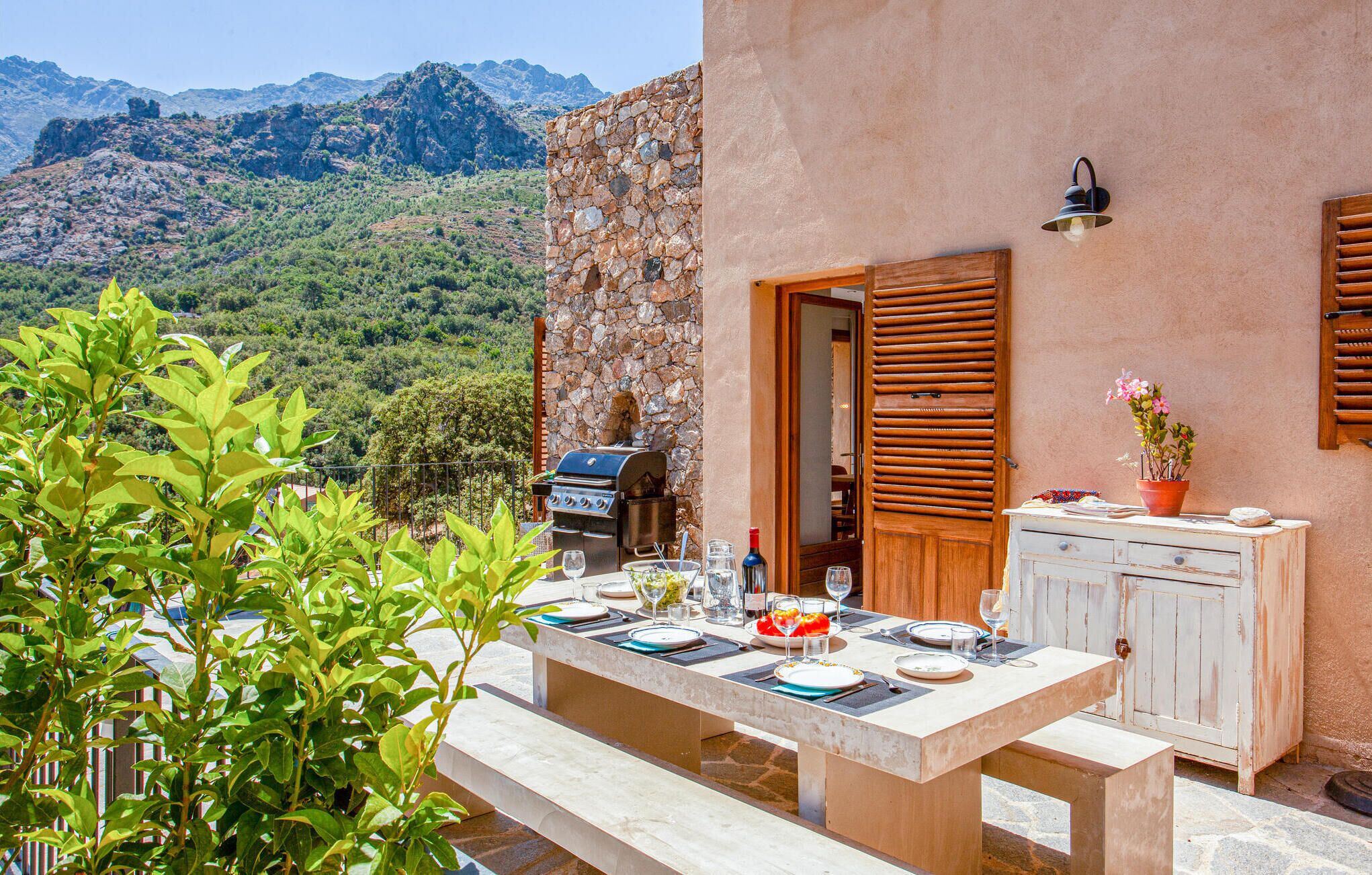 An outdoor dining area with a stone wall and wooden shutters, overlooking a mountainous landscape.