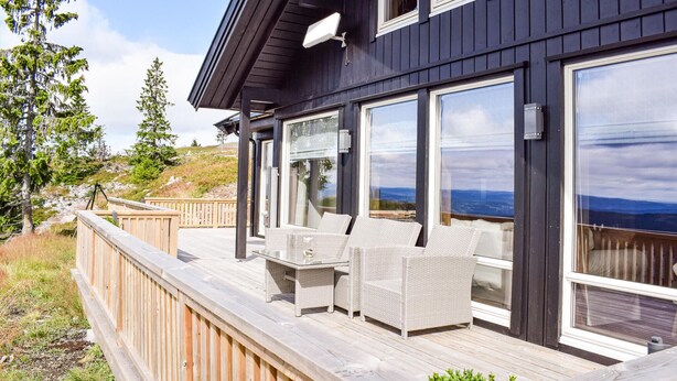 Una terraza de madera con mobiliario exterior, ventanas amplias y vistas del paisaje.