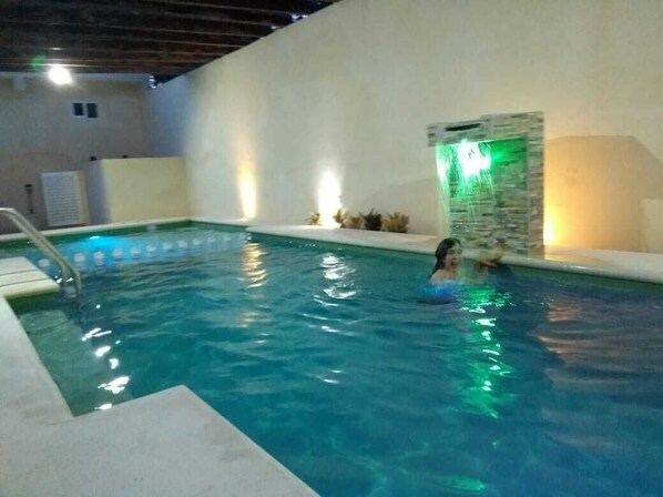 Swimmers can enjoy the pool surrounded by a deck with a ladder and a decorative waterfall feature.