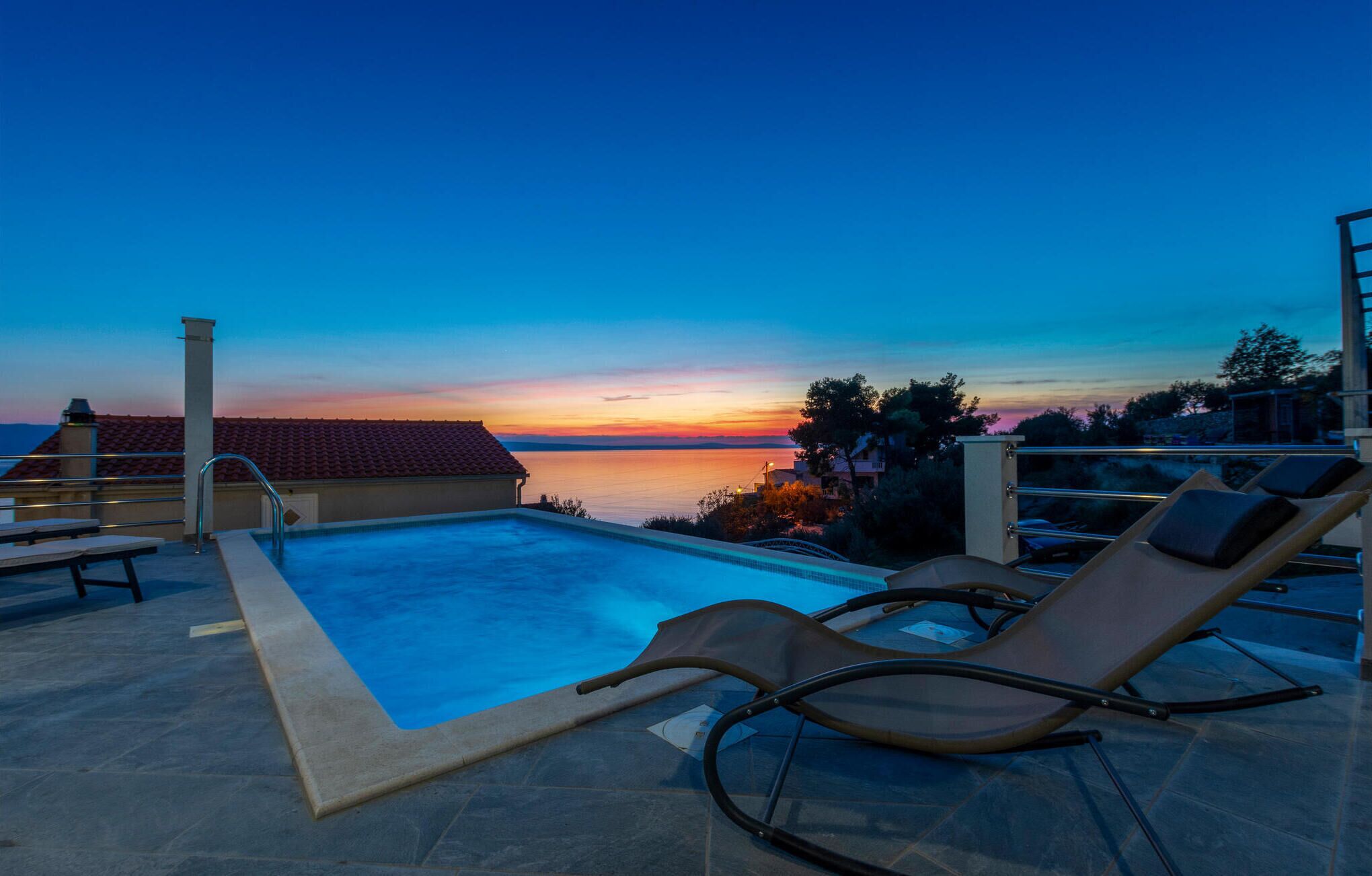 Ein Poolbereich mit Liegestühlen, einer gefliesten Terrasse und Blick auf den Sonnenuntergang über dem Wasser.