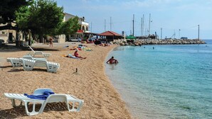 Una playa con sillas de plástico blancas, un edificio con techo rojo y un rompeolas rocoso con barcos.