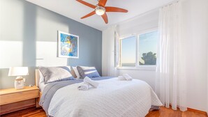 Un dormitorio con cama, mesita de noche, ventilador de techo, ventana con cortinas y un cuadro en la pared.