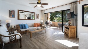 A spacious living room with a wooden floor, a sofa, a coffee table, and a television. There is a balcony with a table and chairs, and a large window providing a view of the outdoors. - Chac Hal Al, Casa Calandria lovely studio steps from the beach. (Puerto Aventuras)