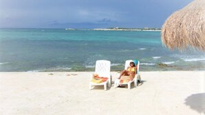 A beach with two white lounge chairs and a straw umbrella. - Chac Hal Al, Casa Calandria lovely studio steps from the beach. (Puerto Aventuras)