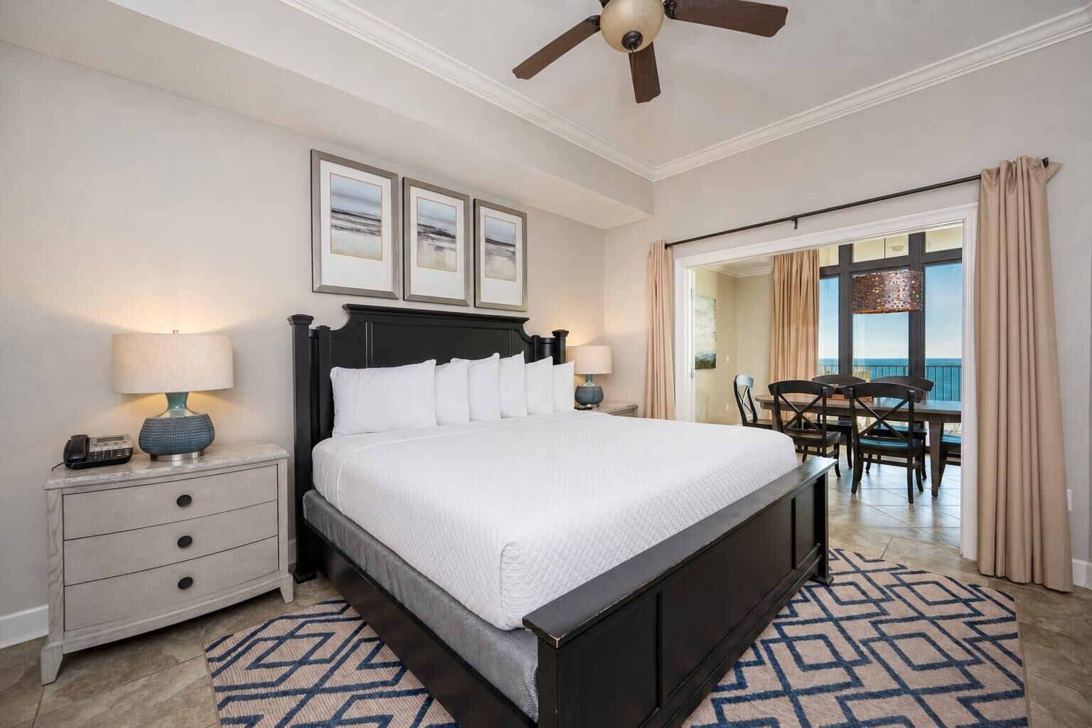 A hotel room with a large bed, bedside lamps, a ceiling fan, a dining area with chairs, and a view through a door.