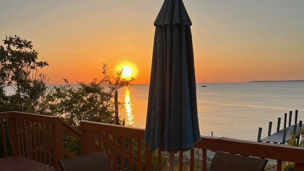 A deck with a table and chairs, overlooking a sunset over the water. - Soundfront with Gorgeous View and Private Pier! (Kill Devil Hills)