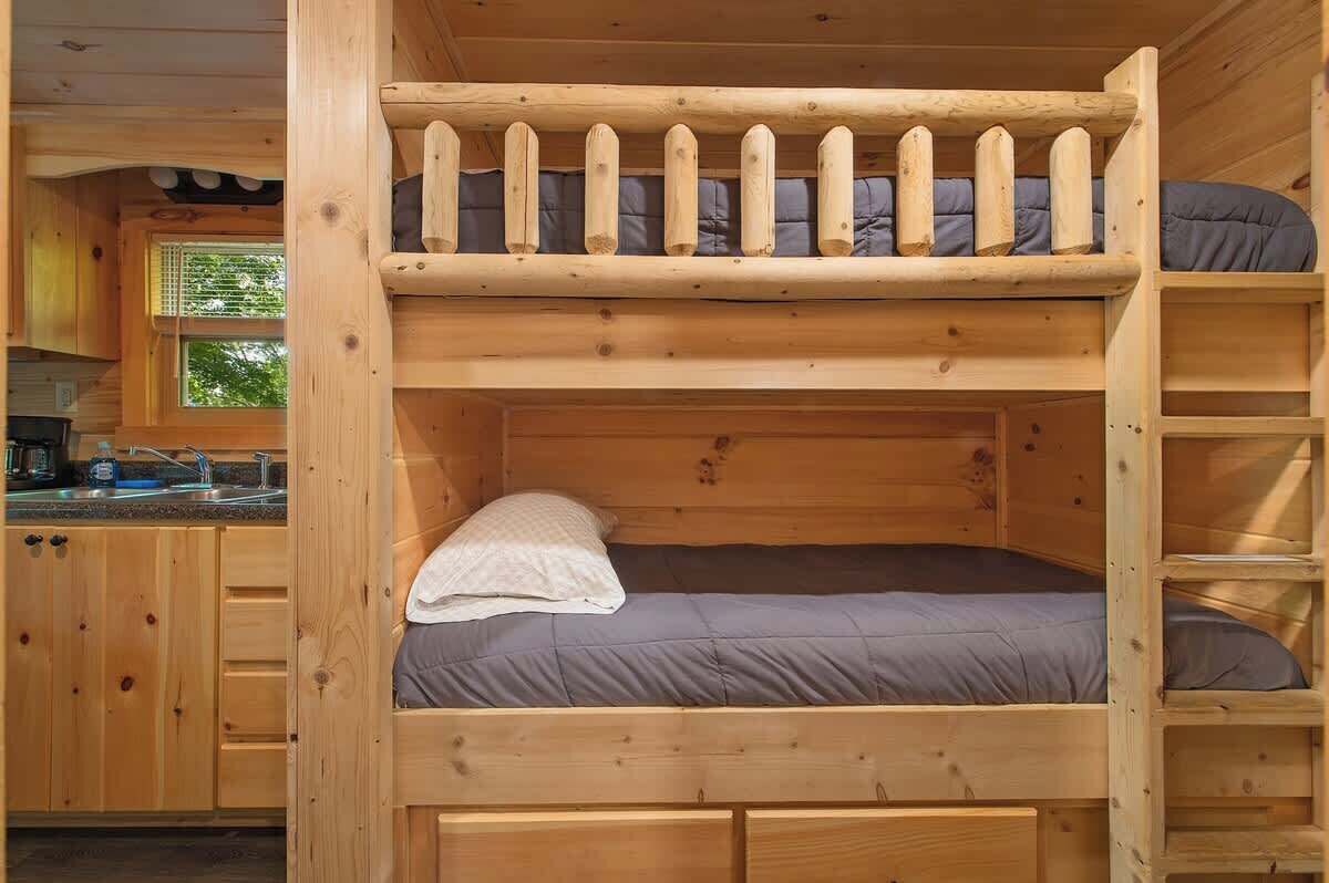 Un lit superposé avec une échelle en bois, un coin cuisine avec des placards et une fenêtre, et un évier.