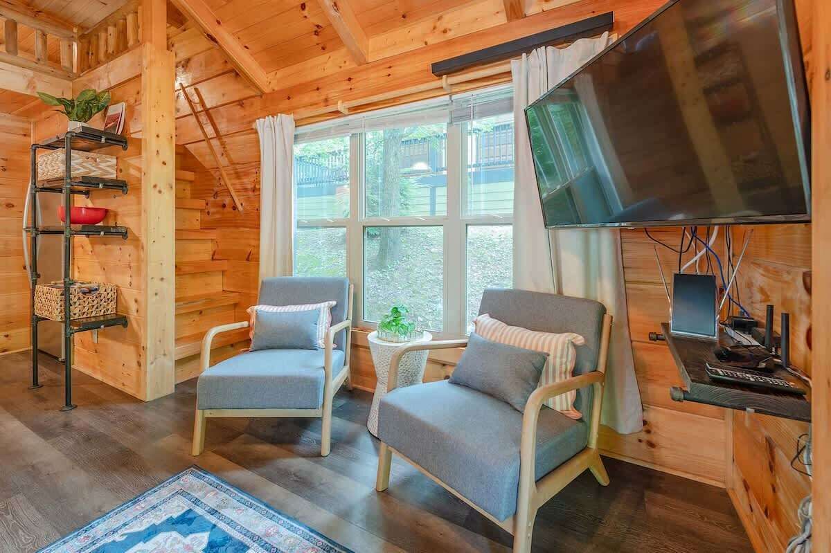 A cozy cabin interior with two grey armchairs, a TV, a shelf with a plant, and a window with curtains.