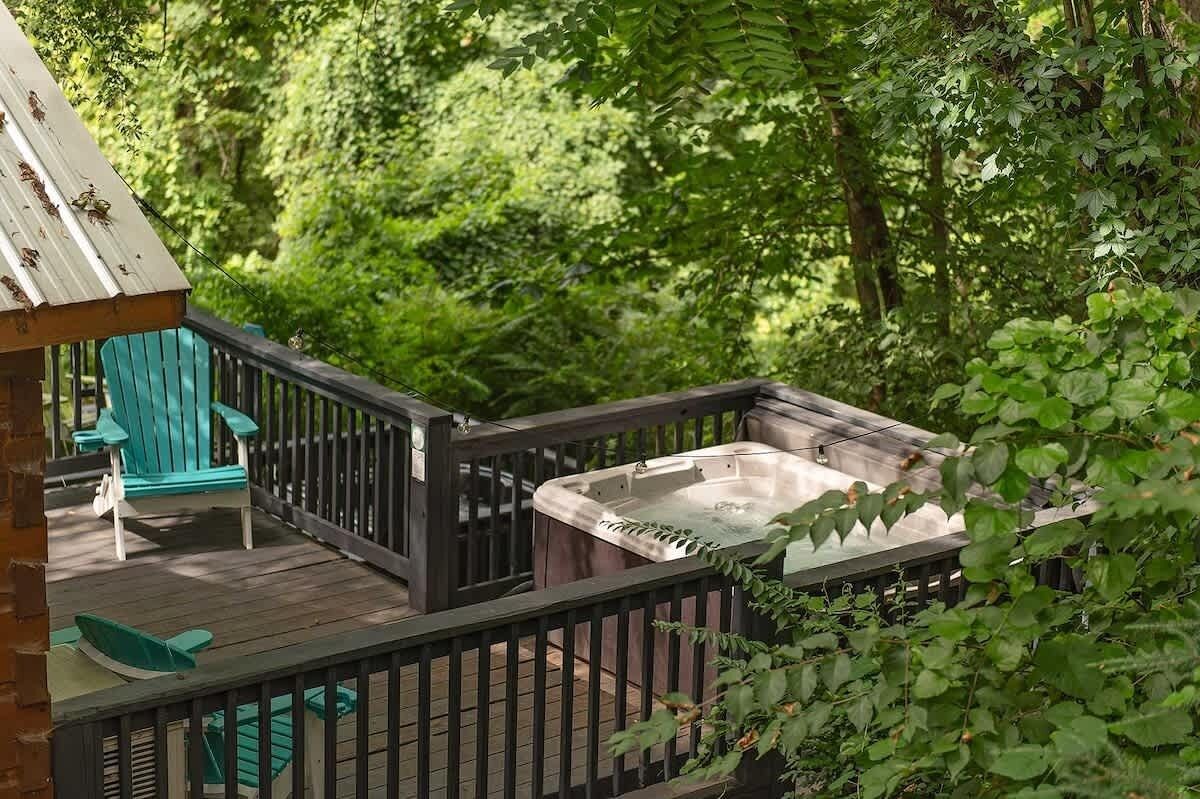 Une terrasse en bois avec un jacuzzi, des fauteuils Adirondack verts et une vue sur une végétation luxuriante.