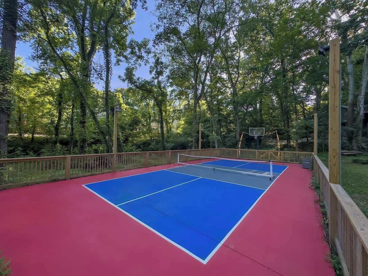 Una cancha de baloncesto al aire libre con superficie roja y azul, rodeada por una valla de madera y árboles.