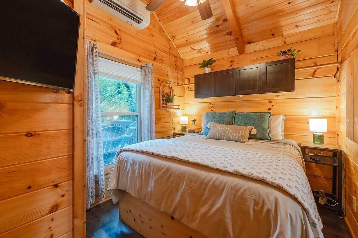 Une chambre dans une cabane en bois, avec un lit, des tables de chevet, un téléviseur fixé au mur et une fenêtre avec des rideaux.