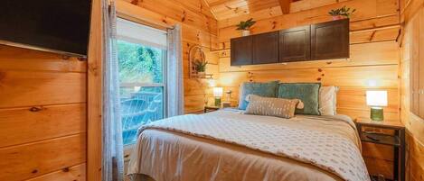 Une chambre dans une cabane en bois, avec un lit, des tables de chevet, un téléviseur fixé au mur et une fenêtre avec des rideaux.