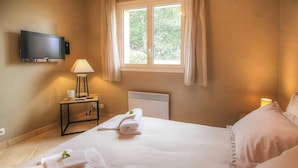 Una habitación de hotel con cama, televisor montado en la pared, ventana con cortinas, mesita de noche con lámpara y radiador.