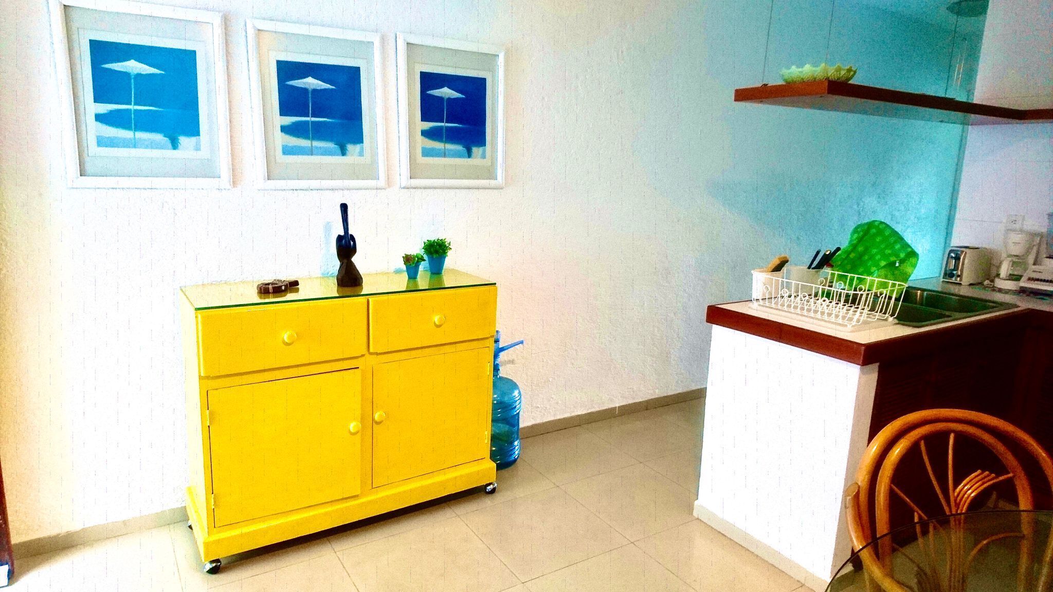 Eine Küche mit einem gelben Schrank, einem blauen Wasserspender und einer weißen Arbeitsplatte mit Spüle. Darüber hängen drei gerahmte Bilder an der Wand.