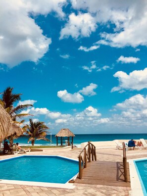 A pool area with a wooden deck, palm trees, and a thatched structure. - Brisas Emerald Apartment - Cancun, Hotel Zone  (Zona Hotelera)
