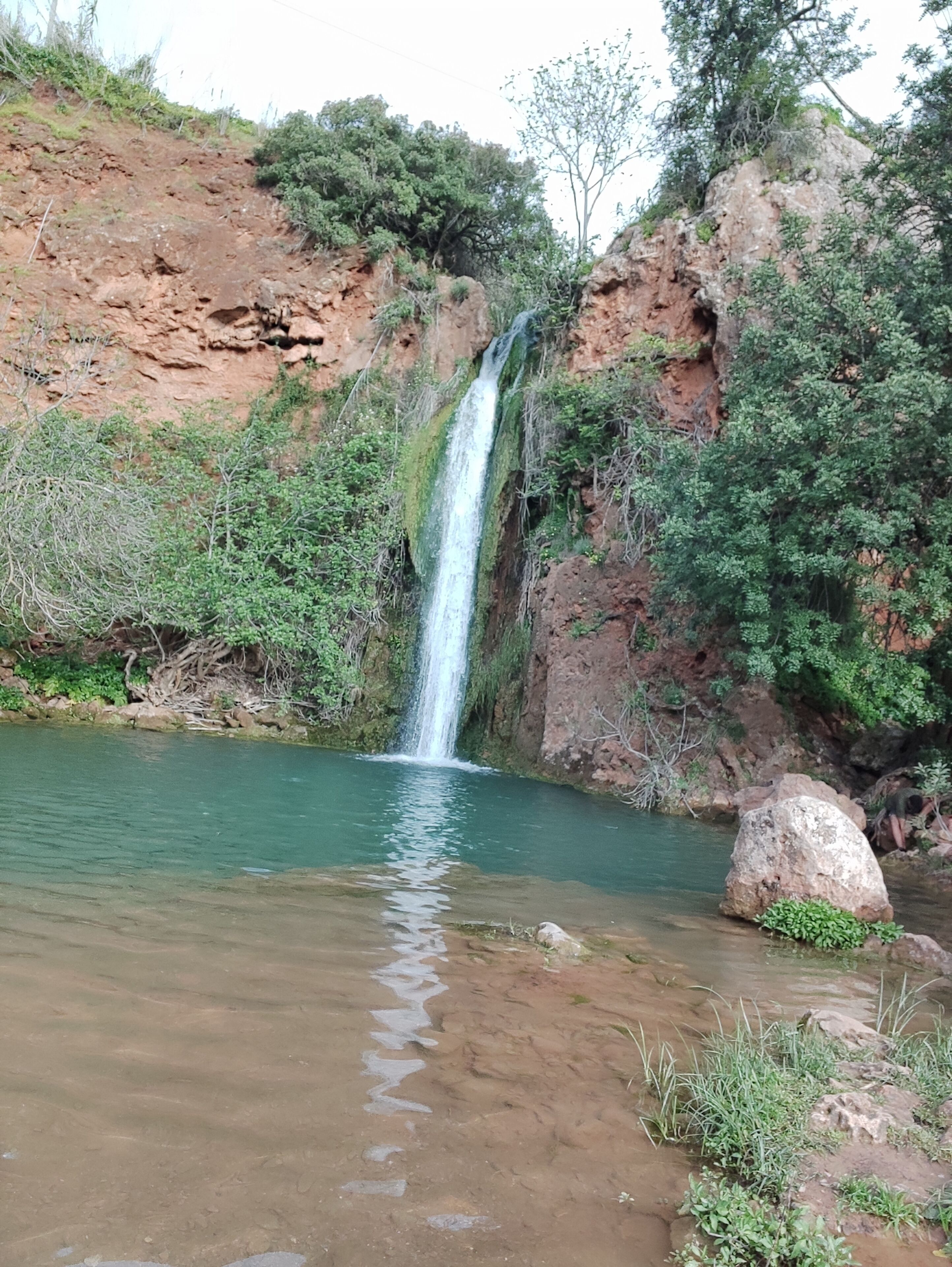 Ein Wasserfall stürzt in ein klares Becken, umgeben von felsigem Gelände und spärlicher Vegetation.