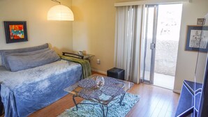 A room with a bed, a glass table with a vase, a lamp, a sliding glass door, and a framed picture on the wall.