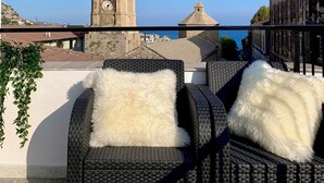 Un balcón con sillas de mimbre y cojines blancos, con vistas a una torre histórica y un paisaje urbano costero.
