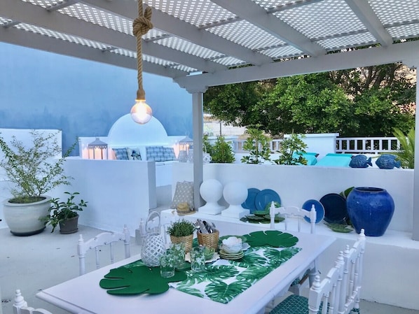 Una terraza en la azotea con una mesa blanca dispuesta para comer, rodeada de plantas en macetas y objetos decorativos.