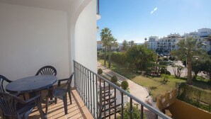 A balcony with a table and chairs, overlooking a well-maintained garden with palm trees and a clear view of the surrounding buildings. - Marina de la Duquesa 225 (Manilva)
