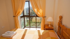 A hotel room with a large window, a bed with a quilt, a bedside table with a lamp, and a wooden headboard. - Marina de la Duquesa 225 (Manilva)