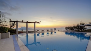 Piscina com pérgola e espreguiçadeiras, rodeada por um jardim ajardinado e vista para um corpo de água.