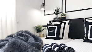 Une chambre moderne avec un lit, des coussins à rayures noires et blanches, un plaid gris moelleux et une table de chevet agrémentée d’une plante et de livres.