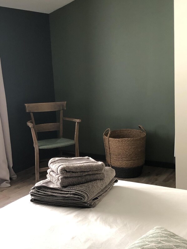 Una cama bien tendida con toallas dobladas, una silla de madera y una cesta tejida frente a una pared verde oscuro.