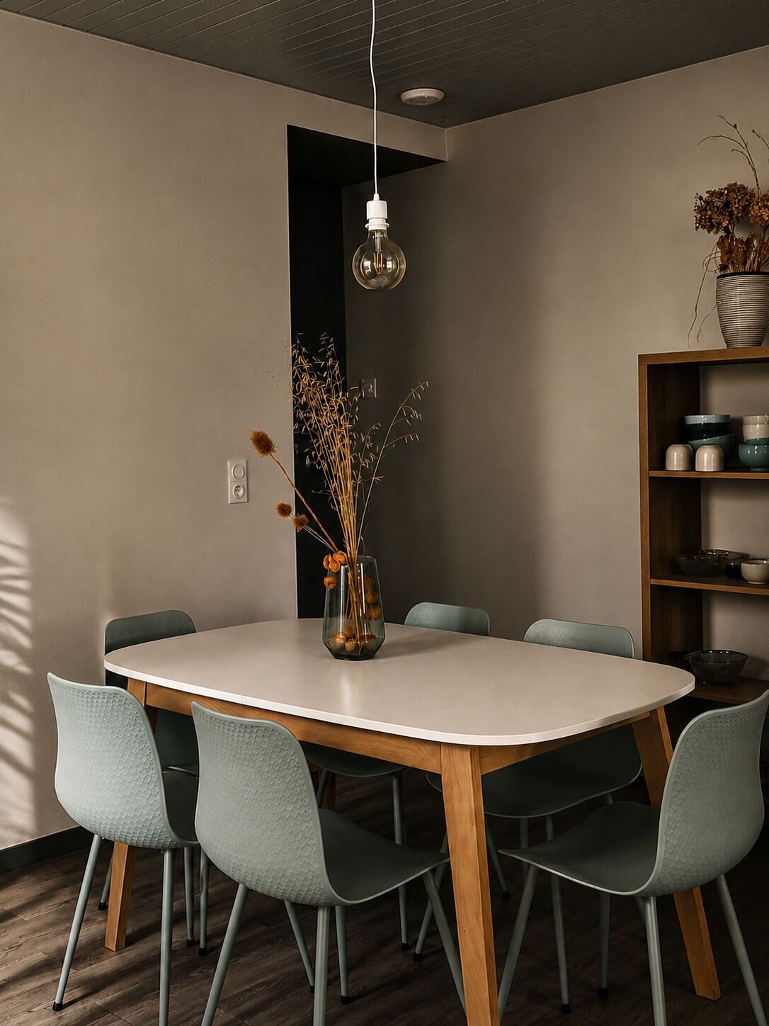 Un comedor moderno con una mesa redonda, seis sillas, un jarrón con plantas secas y una estantería con diversos objetos.