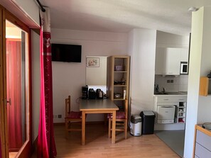 A compact kitchen with a dining area, a television mounted on the wall, and a trash bin.