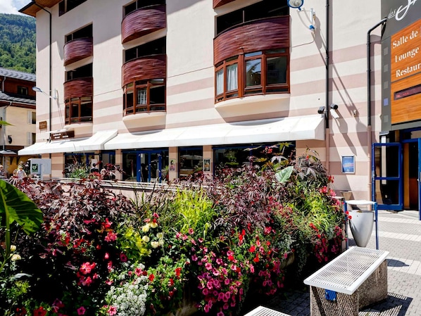 Ein Hotel mit überdachter Außenbereich mit Sitzgelegenheiten, einem Schild für 'Salle de Loungue Restaurant' und verschiedenen Pflanzen und Blumen.