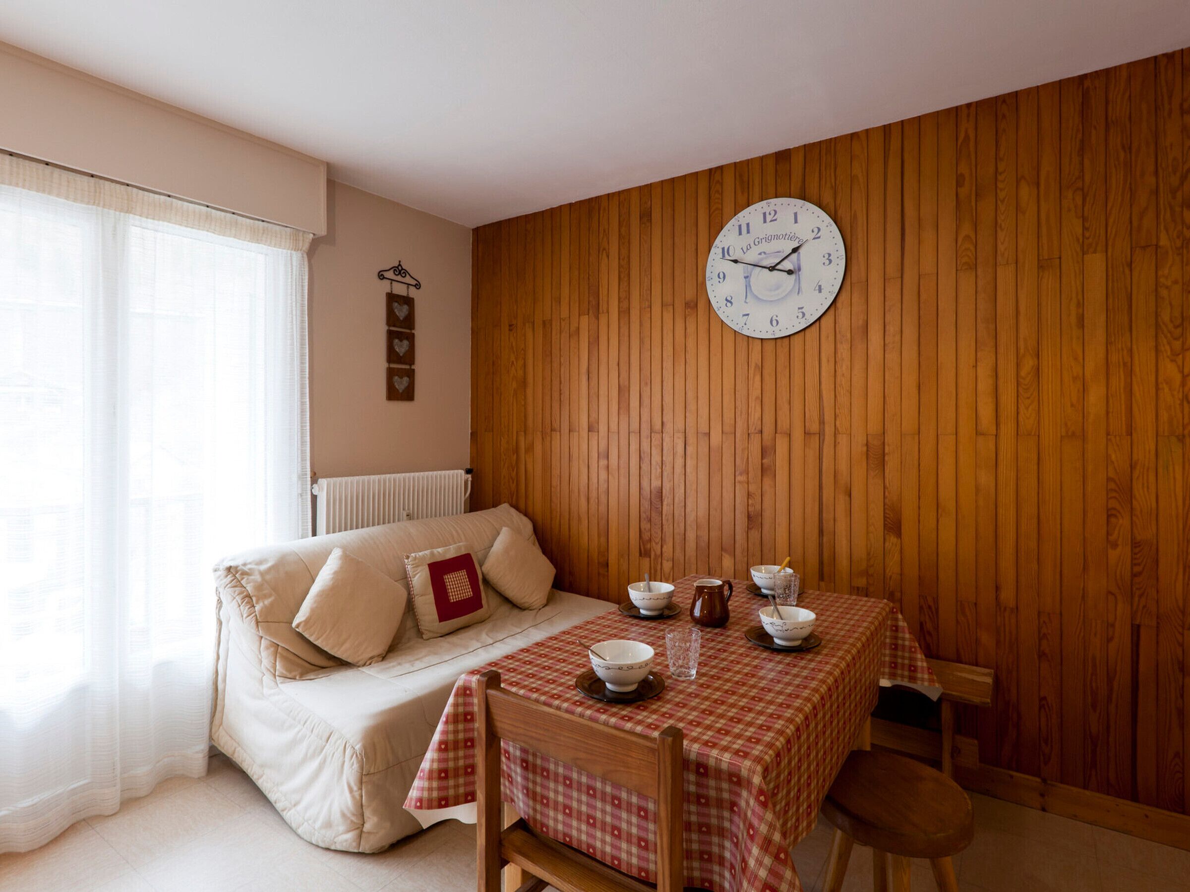 Una habitación acogedora con pared de madera, un reloj de pared redondo, un sofá con cojines, una mesa con mantel a cuadros y una ventana con cortinas translúcidas.