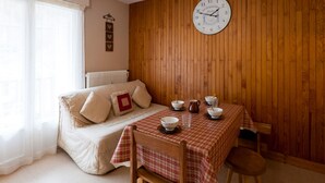 A cozy room with a wooden wall, a round wall clock, a sofa with cushions, a table with a checkered tablecloth, and a window with sheer curtains. - Studio for 5, Central, South Balcony, Elevator - Brides-les-Bains (Brides-les-Bains)