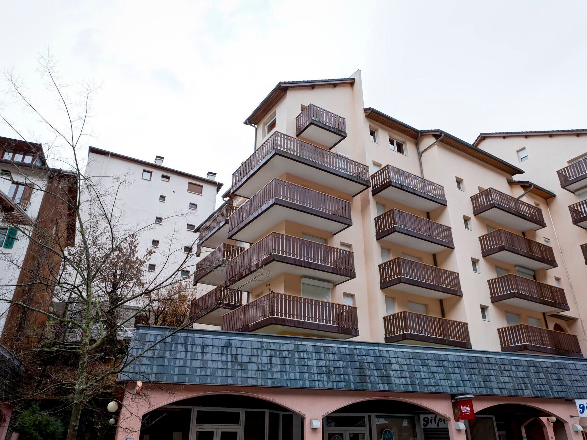 Un edificio residencial de varios pisos con balcones, una tienda con un letrero y un árbol en primer plano.