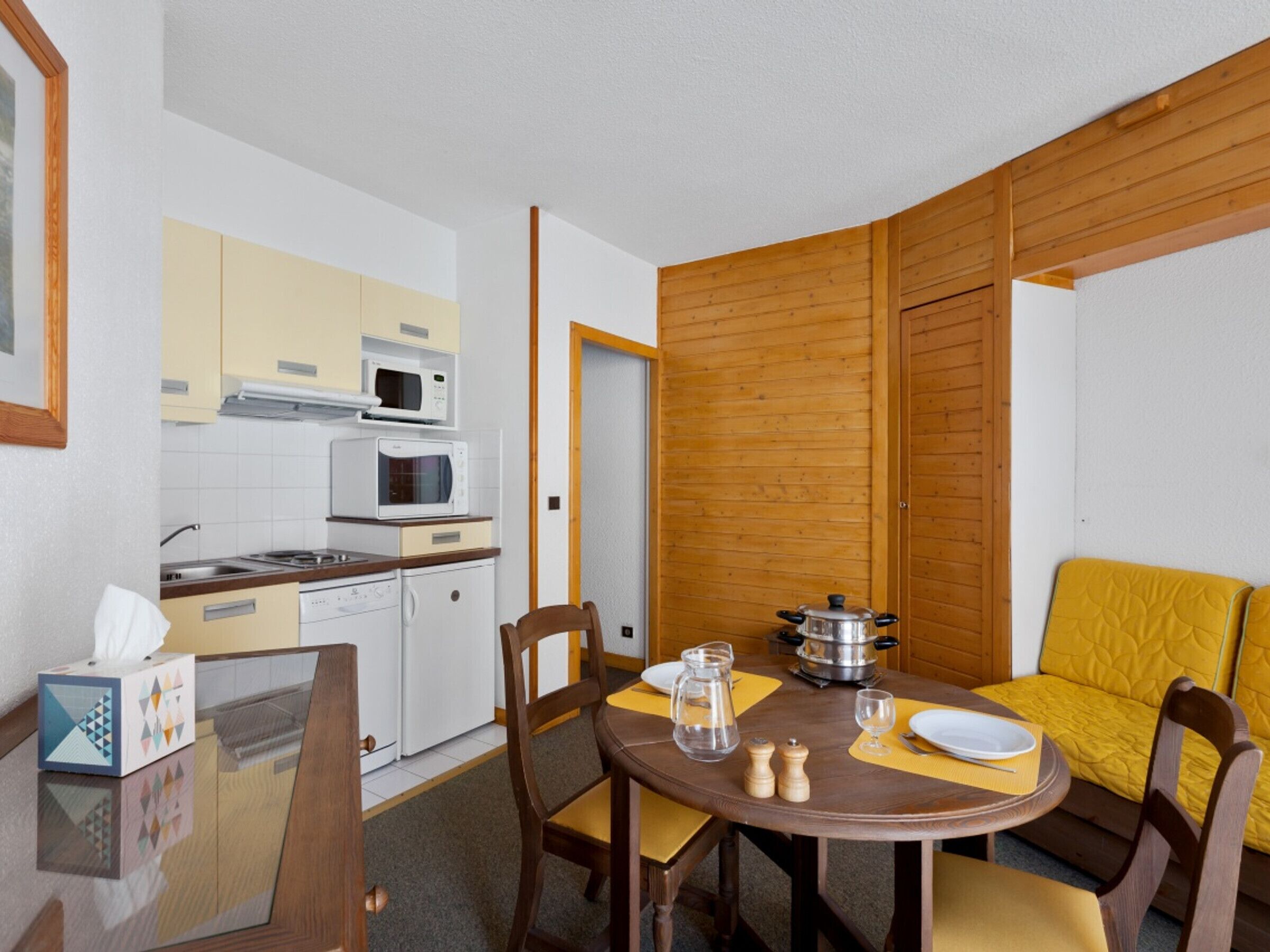 Una cocina compacta con microondas, estufa y pileta, una mesa de comedor con sillas y un sofá amarillo.