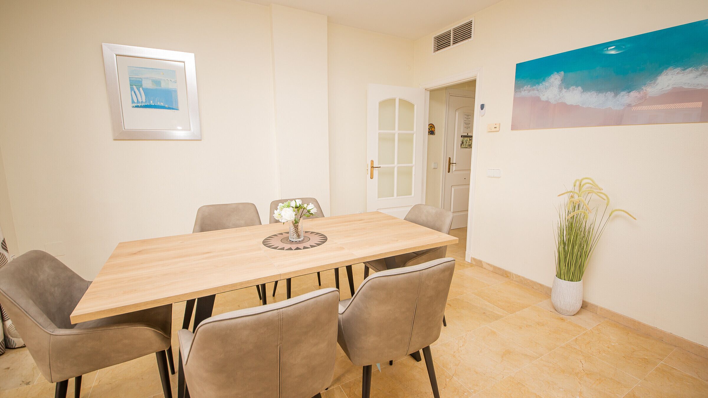 A dining area with a wooden table, six chairs, a vase with flowers, and two wall-mounted paintings.