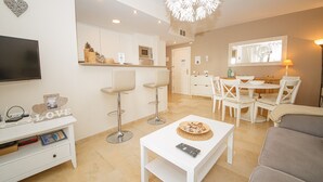 Uma sala de estar moderna com televisão de tela plana, móveis brancos, área de jantar com mesa e cadeiras e cozinha com micro-ondas e forno.
