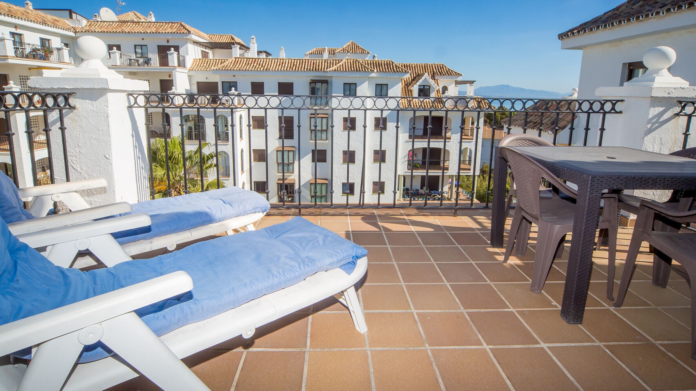 Una terraza con dos tumbonas, una mesa y vistas a un edificio blanco con balcones.